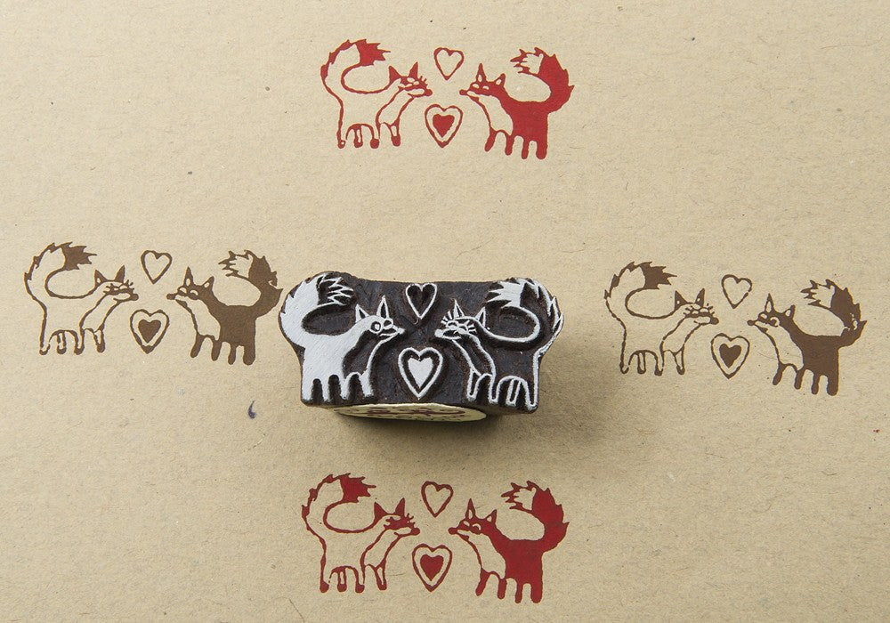 A hand-carved wooden printing block with a design of foxes in love, placed on a beige background with heart shapes.