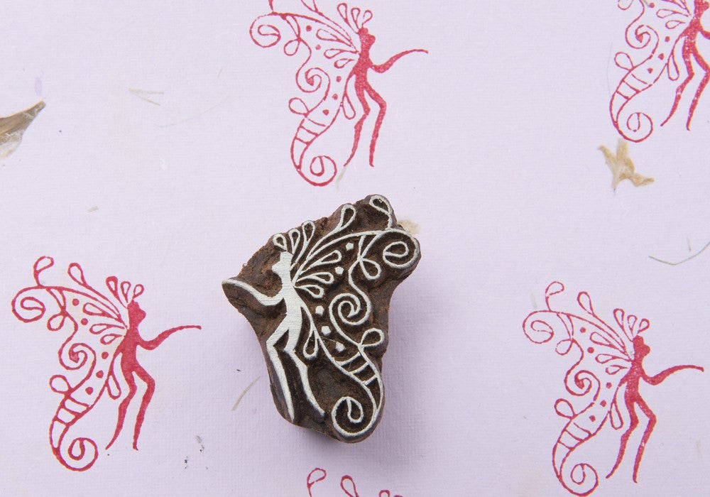 A rectangular wood block stamp with a fairy design, predominantly brown with white outlines, placed on a pink background with red dots.