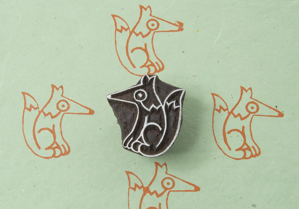 A hand-carved wooden printing block of a fox, with an orange and white color scheme, placed on a green background with fox silhouettes in the background.